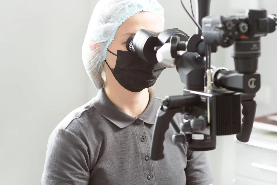 A dentist wearing a mask uses a microscope in a dental clinic while treating a patient.